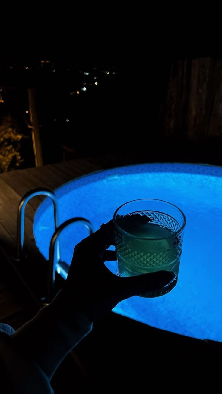 a person holding a glass in front of a swimming pool at night with the lights on