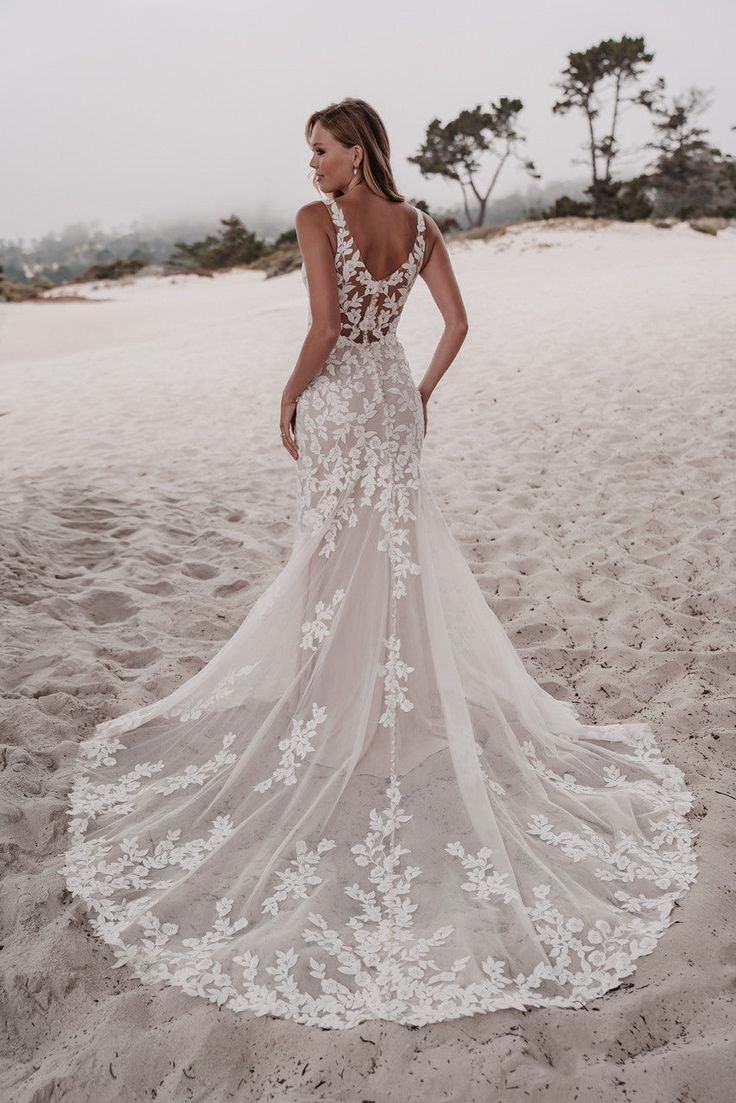 a woman standing on top of a sandy beach wearing a wedding dress with white flowers