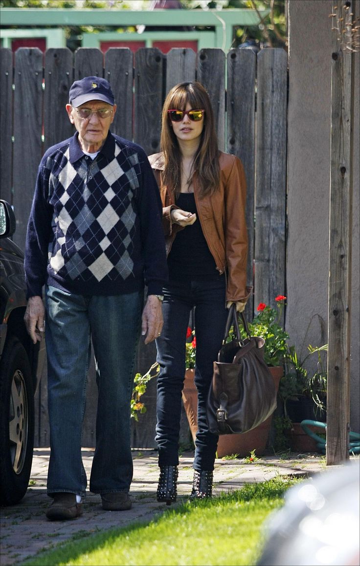 an older man and young woman walking down the sidewalk in front of a fenced area