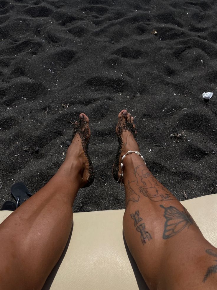 a person's legs with tattoos on them sitting in the sand at the beach