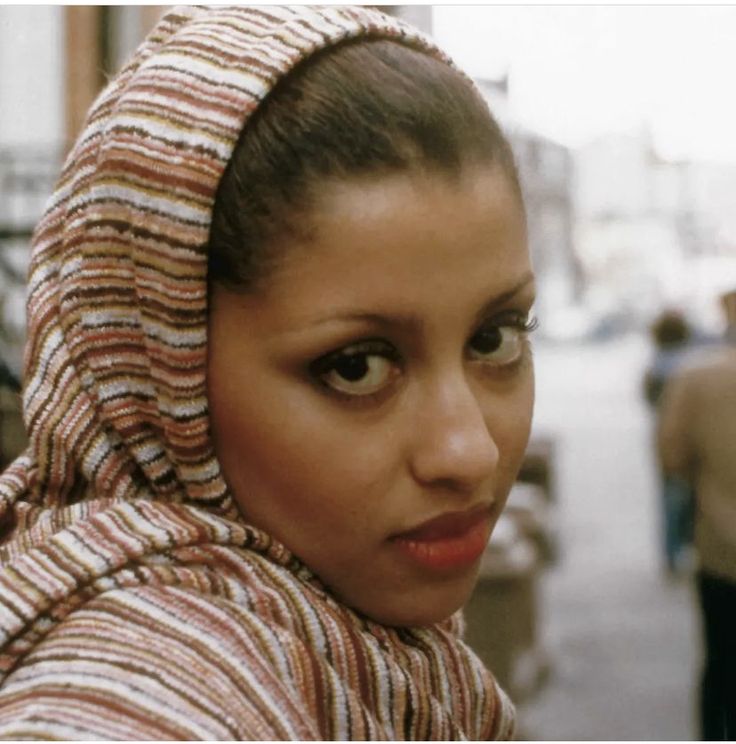 a woman with a scarf on her head is looking at the camera while people are walking in the background