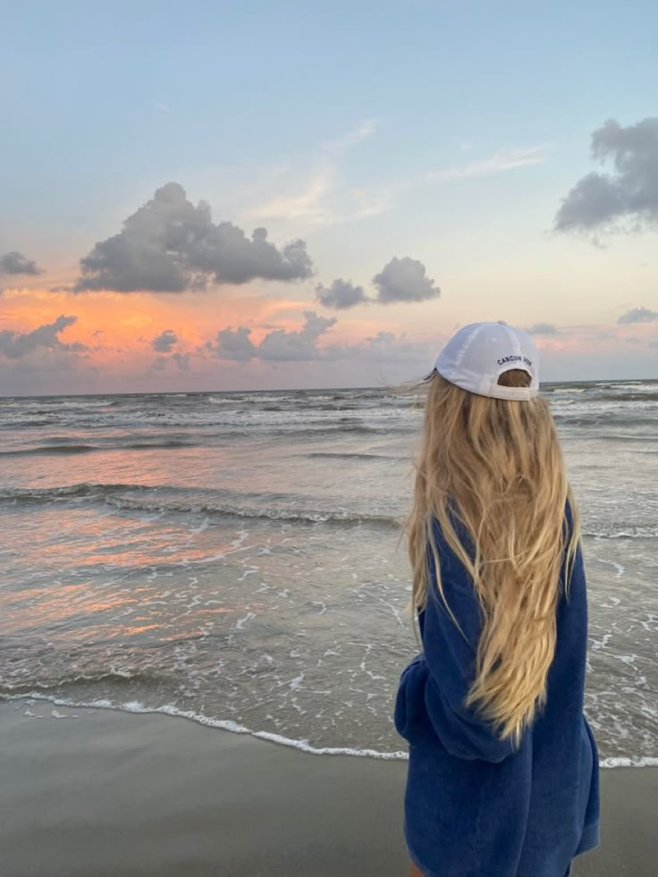 a woman standing on top of a sandy beach next to the ocean at sunset with clouds in the sky