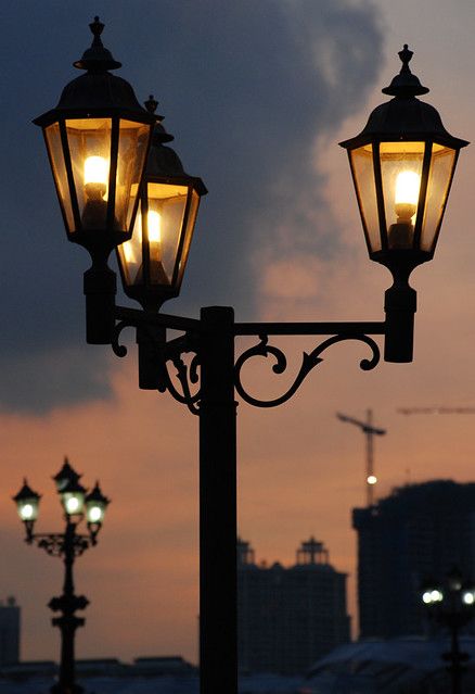 two street lamps are lit up against the evening sky in front of a cityscape