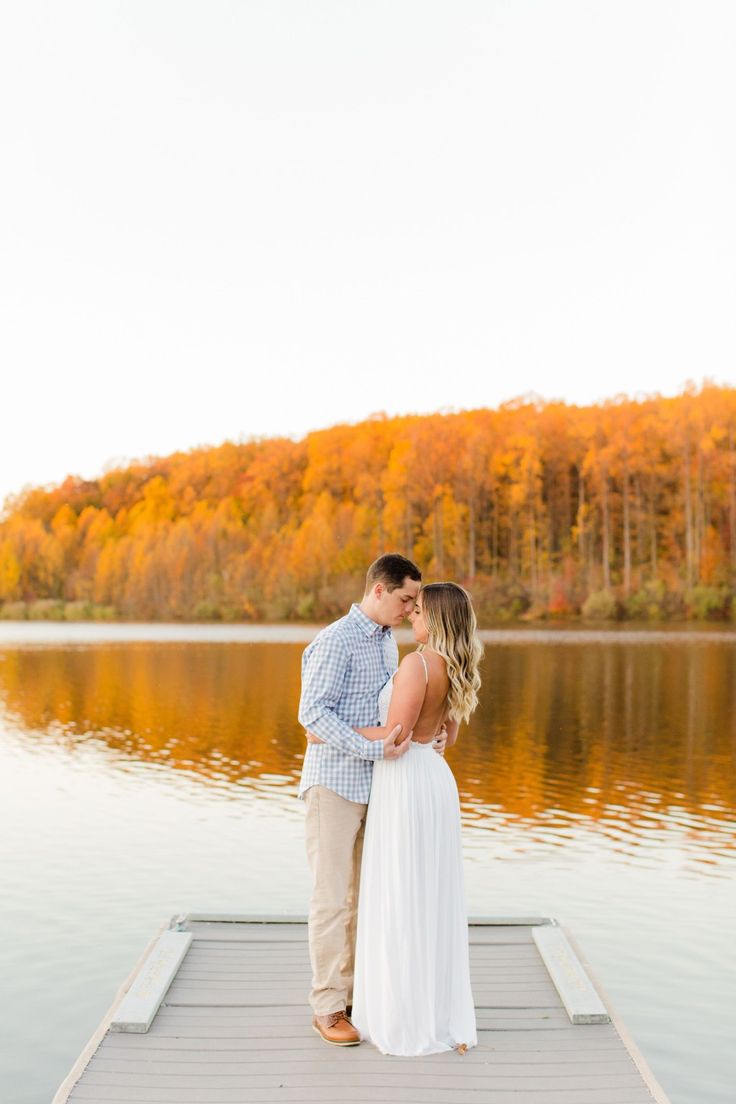 an engaged couple standing on a dock in front of a lake surrounded by fall foliage