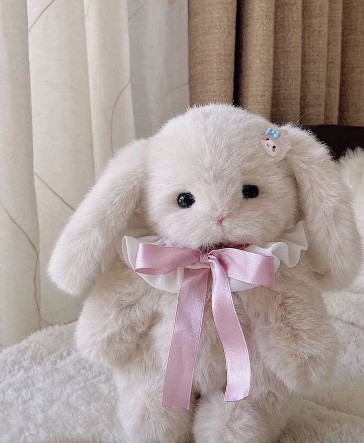 a white stuffed rabbit with a pink ribbon around its neck sitting on top of a bed