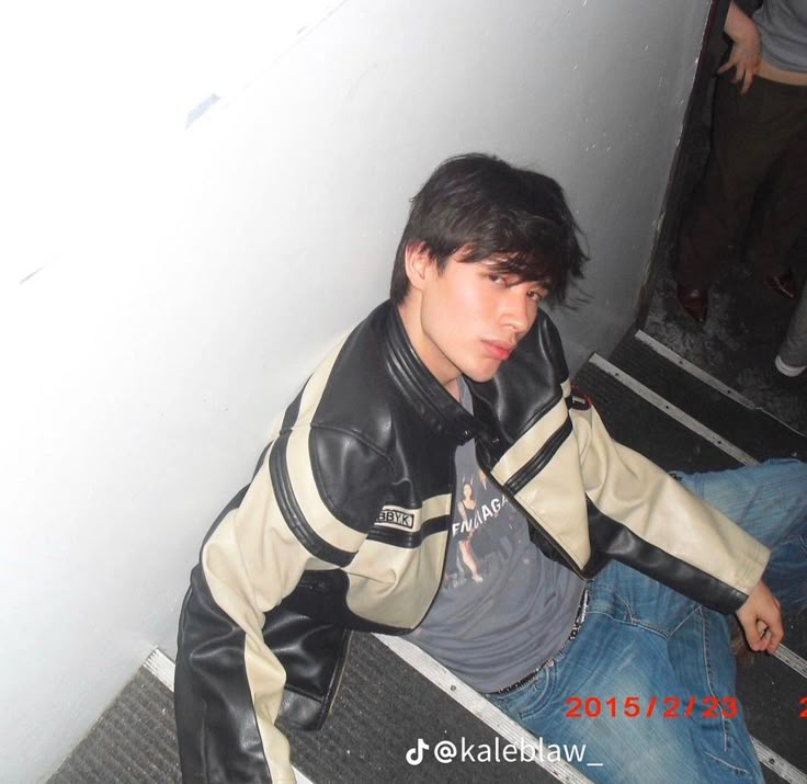 a young man sitting on the ground wearing a leather jacket and jeans, leaning against a wall