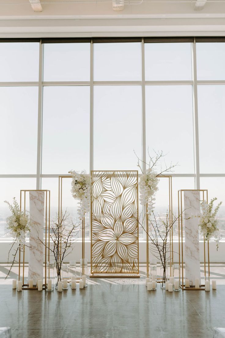 an elegant wedding setup with white flowers and gold accents in front of large glass windows