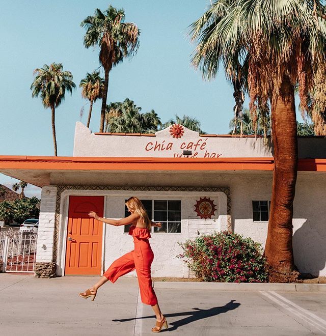 a woman in an orange jumpsuit is dancing outside of a building with palm trees