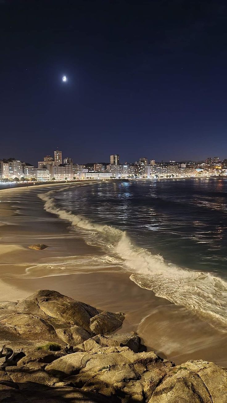 the beach is lit up at night with city lights in the background and waves crashing on the shore