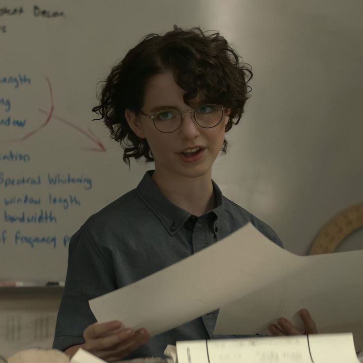 a woman in glasses holding papers while standing next to a whiteboard with writing on it