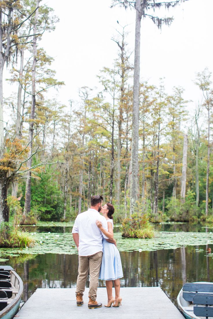 a man and woman kissing on a dock in front of a lake surrounded by trees