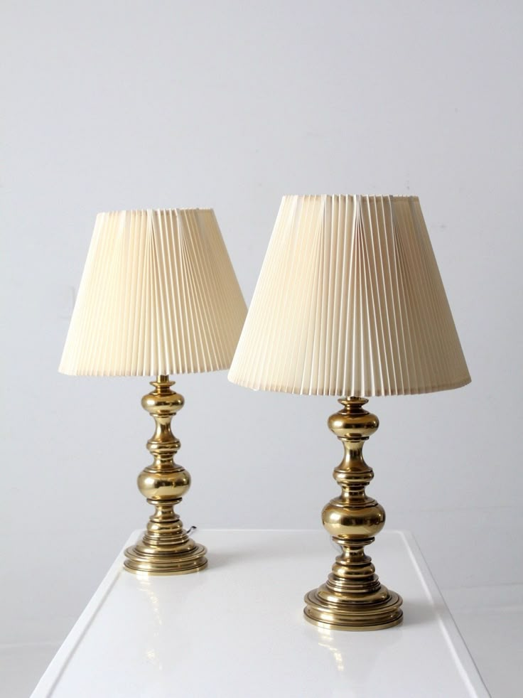 two golden lamps sitting on top of a white table next to each other, one with a beige shade