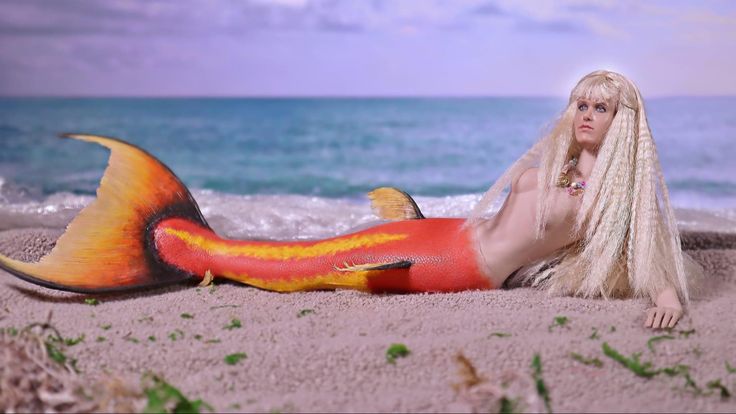 a woman with long blonde hair laying on the beach next to an orange and yellow mermaid tail