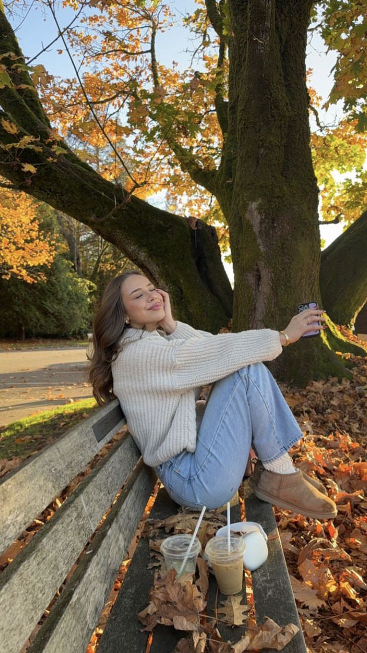 a woman sitting on top of a wooden bench next to a leaf covered tree and holding a cup