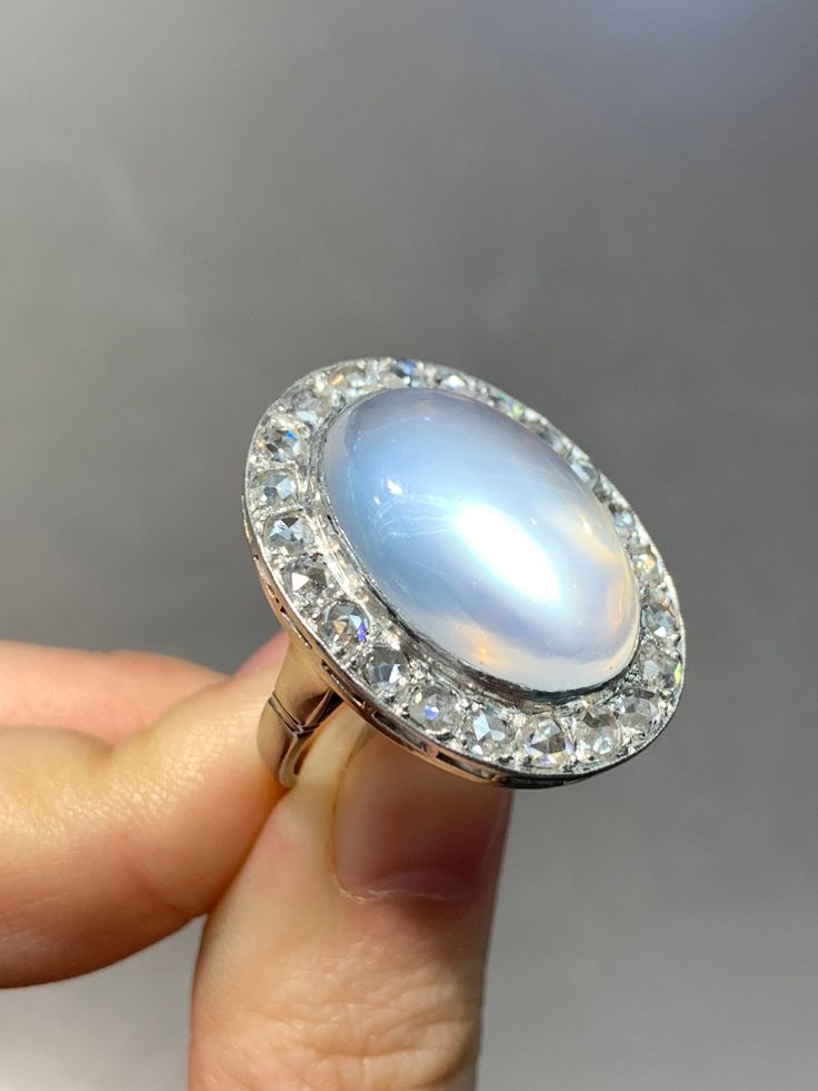 "A dreamy glowing moonstone cabochon ring measuring 2.7 cm by 2.1 cm, encrusted with approximately 1.8 carats of glimmering chunky icy-white rose cut diamonds. The moonstone shines out a eye-catching luminous blue adularescense. Currently measures a size 6. Ring Face Measurement: 2.7 cm by 2.1 cm Measurement of Moonstone: 2.0 by 1.1 cm Approximate Carat Weight of Diamond: 1.8 Carats Gold Purity: 14K Markings: \"14K\" Ring Size: 6.75 (Resizable)" Faceted Moonstone Ring, Opal Diamond Ring Oval Cabochon, Diamond Opal Ring Oval Cabochon, White Moonstone Ring With Rose Cut Diamonds, Luxury Moonstone Gemstone Ring For Anniversary, Diamond White Moonstone Ring With Rose Cut Diamonds, Antique Oval Moonstone Ring, Elegant Domed Moonstone Gemstone Ring, Luxury Round Moonstone Gemstone Ring