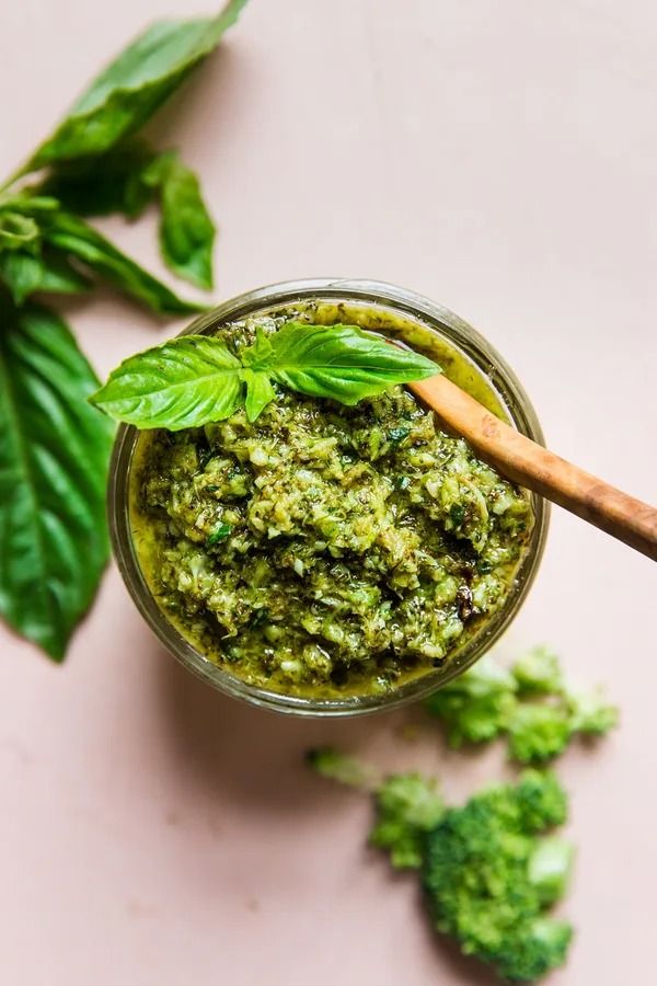 a jar filled with green pesto next to broccoli and leaves on a pink surface