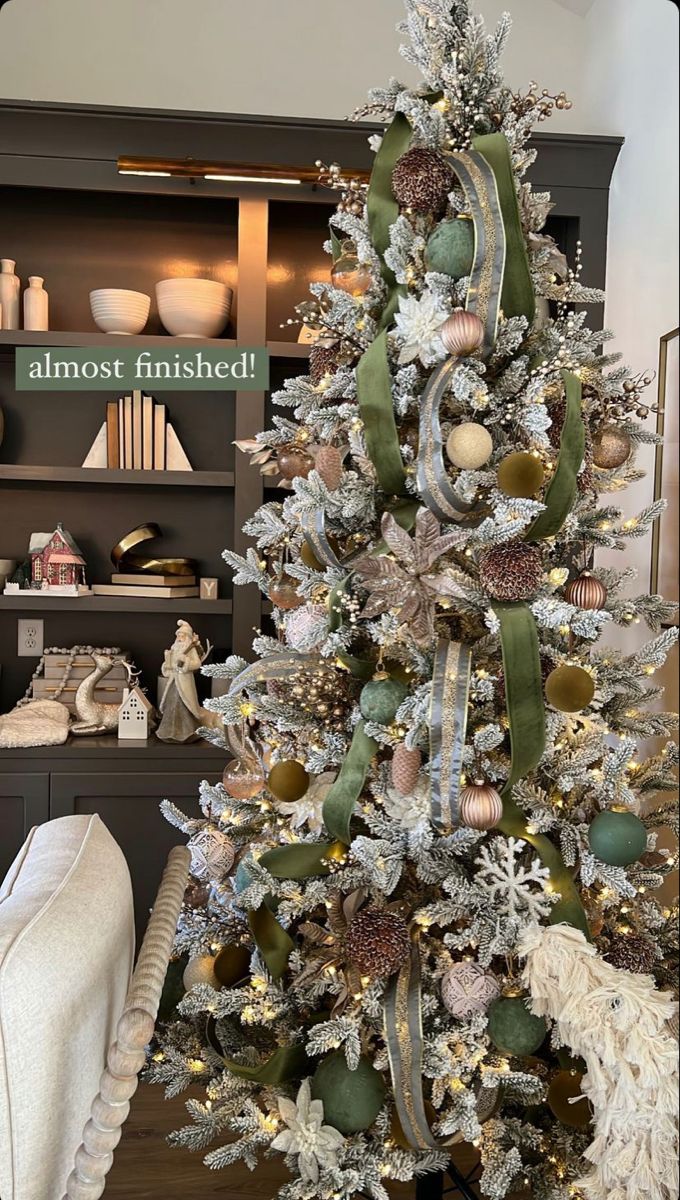 a white christmas tree with green and gold ornaments on it's branches, in front of a bookcase