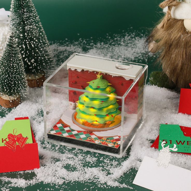 a small christmas tree in a clear box surrounded by cards and snow on the ground