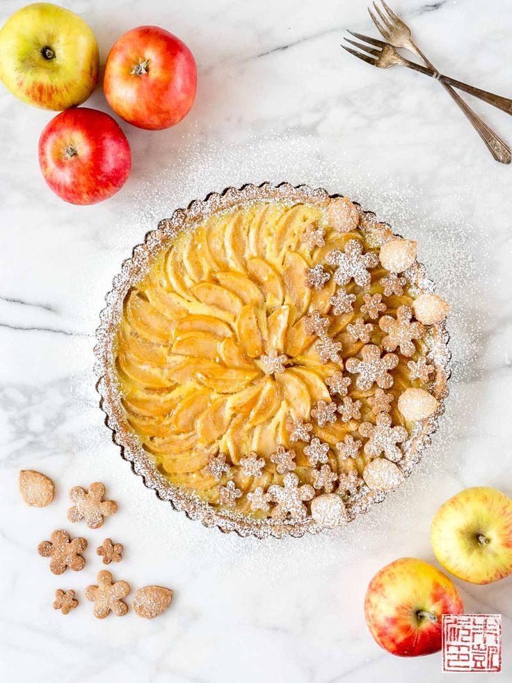 an apple pie with powdered sugar on top next to some apples and spoons