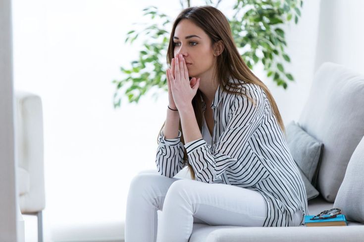 a woman sitting on a couch with her hands clasped to her face and looking away from the camera