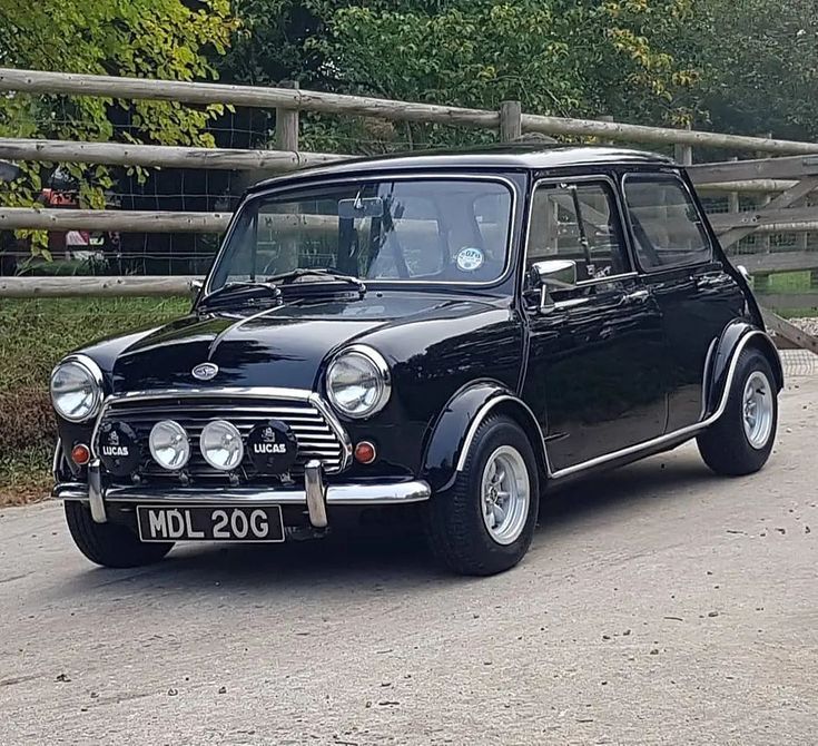 an old black mini car is parked in front of a wooden fence and some trees