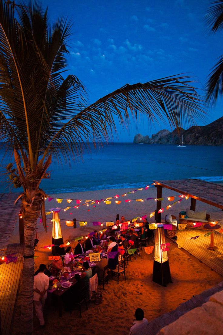 an outdoor dining area on the beach at night with palm trees and lights in the background