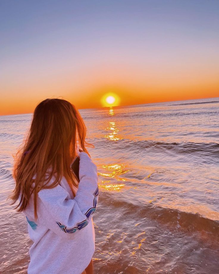 a girl standing on the beach watching the sun rise over the ocean with her hair blowing in the wind