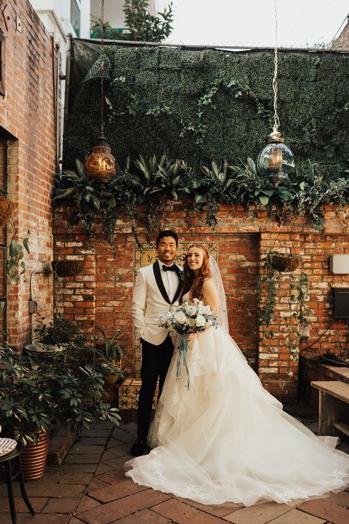 a bride and groom standing in front of a brick wall with greenery on it