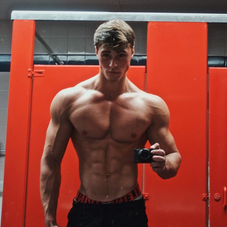 a shirtless young man holding a camera in front of a red wall with his hands on his hips