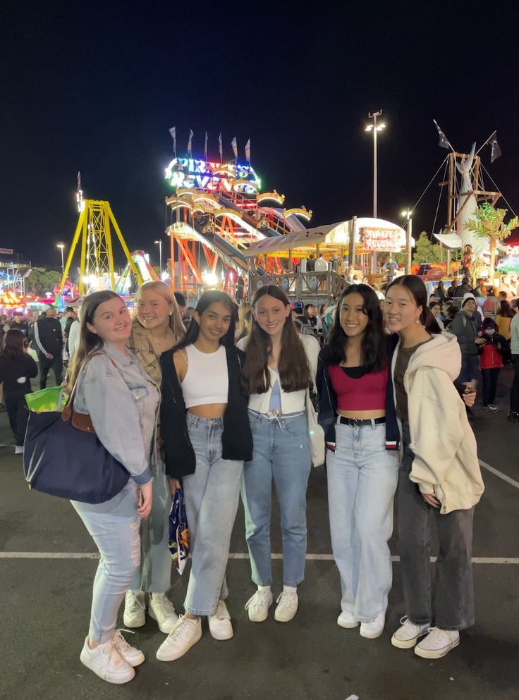 four girls are standing in front of an amusement park at night with ferris wheel in the background