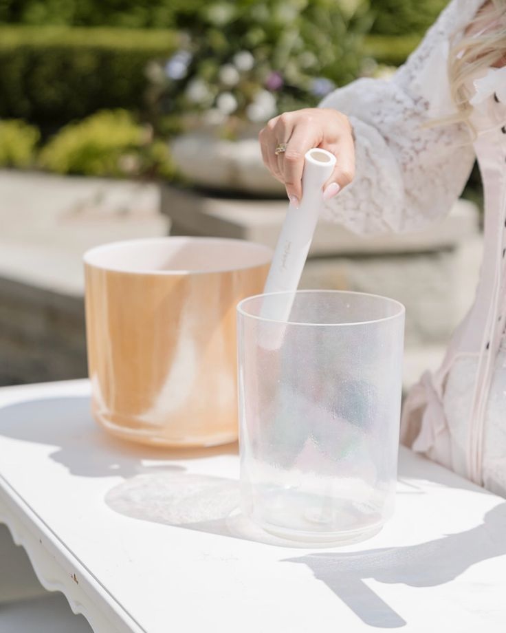 a woman pouring something into a glass on top of a white table with two cups in the background