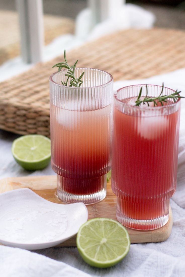 two glasses filled with red liquid sitting on top of a wooden cutting board next to limes