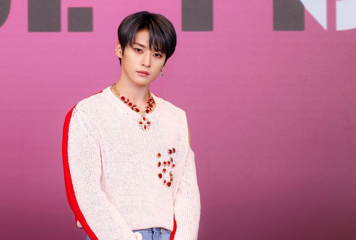 a young man standing in front of a pink wall wearing a white sweater and blue jeans