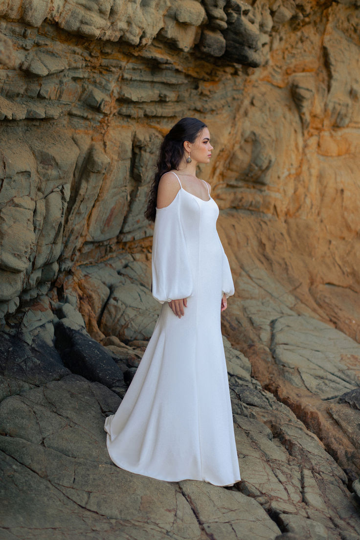 a woman in a white dress standing on rocks near the ocean with her arms behind her back