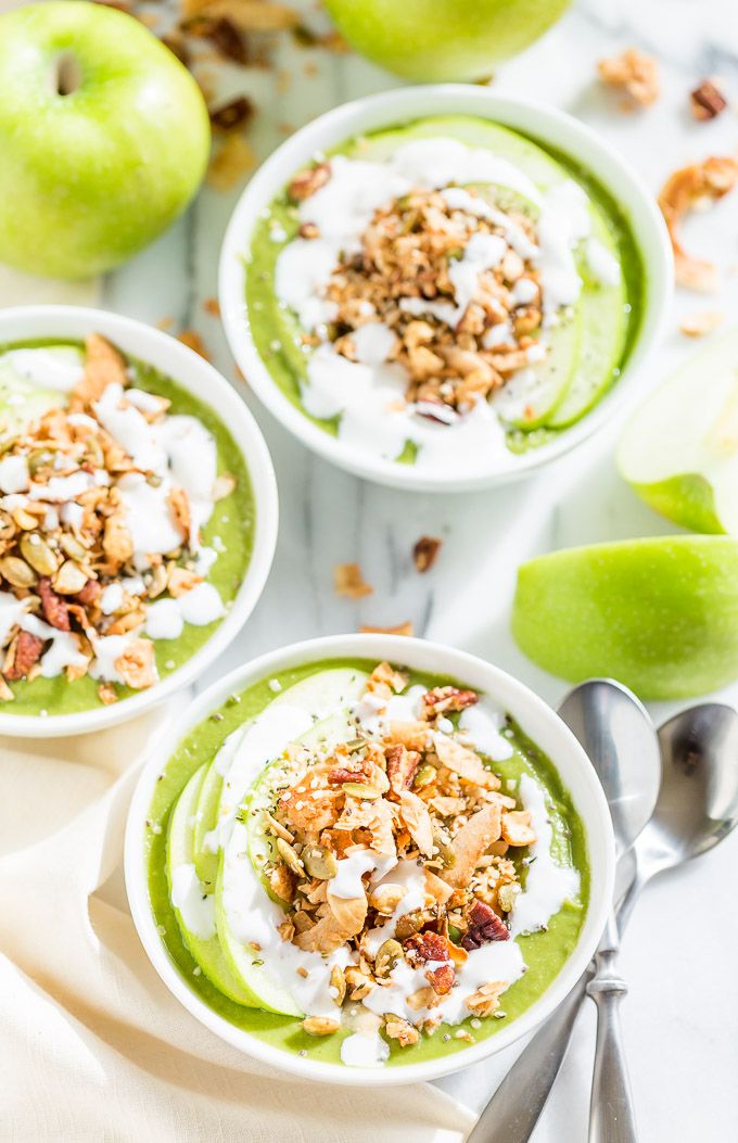 three bowls filled with green smoothie topped with nuts and yogurt next to an apple