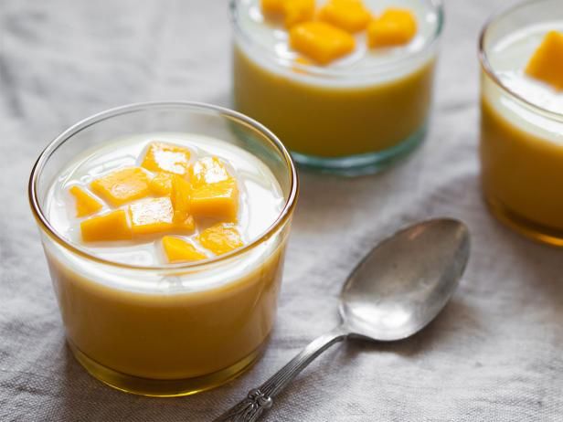 two small bowls filled with pudding and sliced mangoes next to spoons on a table