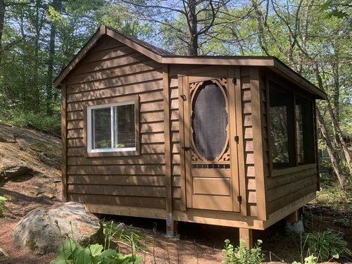 a small wooden cabin in the woods surrounded by trees and rocks, with a screen door