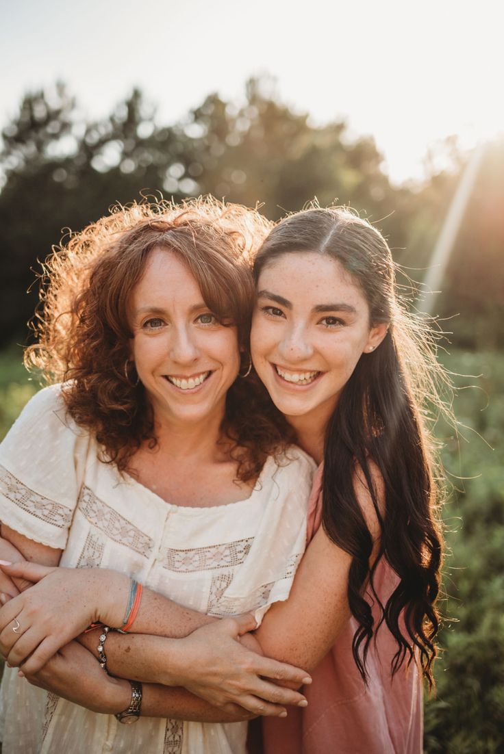 two women hugging each other in front of some trees and bushes with the sun shining on them