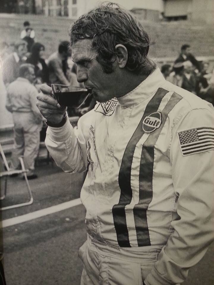 a man drinking from a glass while standing on the side of a road with other people behind him