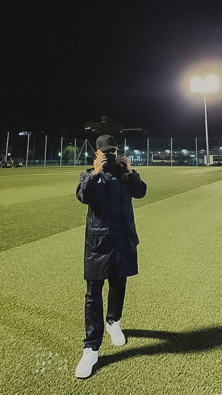 a man standing on top of a field holding a cell phone to his ear while wearing a black jacket