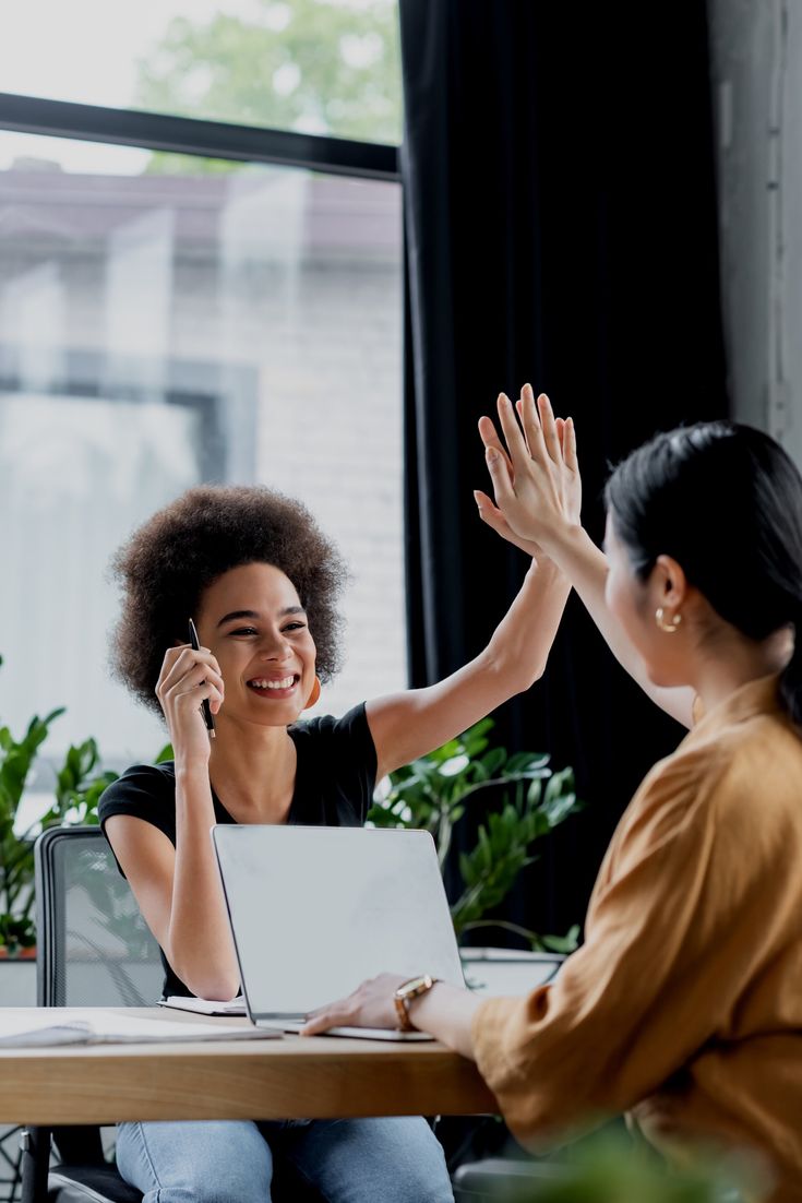 two women sitting at a table with their hands in the air and one woman on her laptop