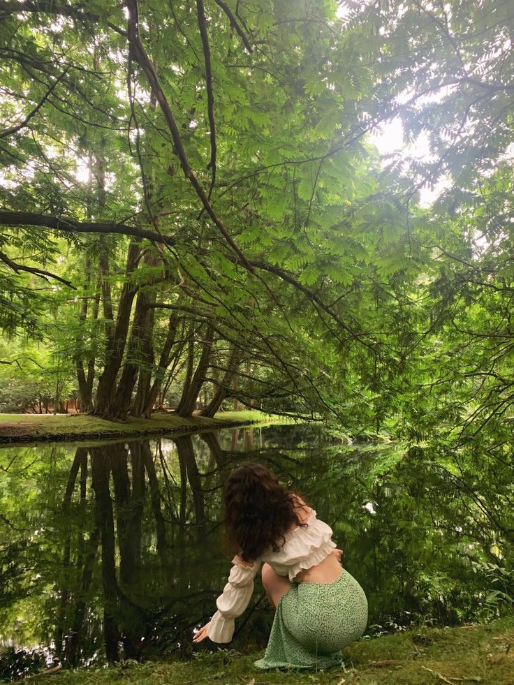 a woman kneeling on the ground next to a pond in front of trees and grass