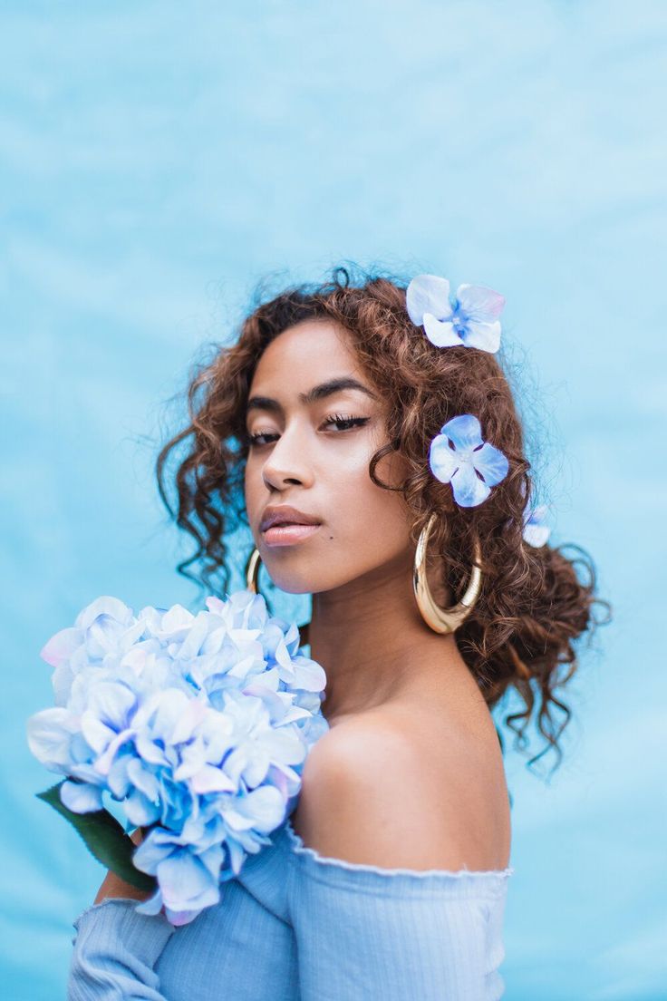 a woman with curly hair and blue flowers in her hair is holding a bouquet of hydrangeas