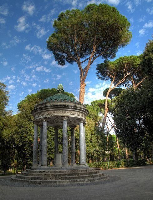 a gazebo in the middle of a park surrounded by trees and bushes, under a blue sky with wispy clouds