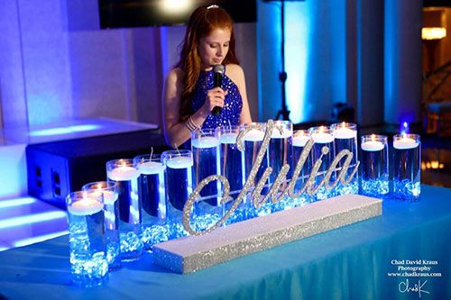 a woman standing in front of a table with many glasses on it and candles behind her