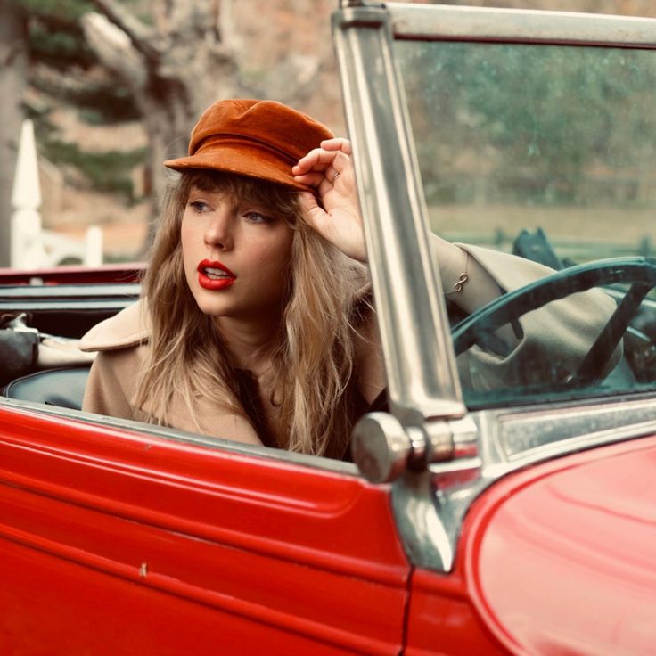 a woman sitting in the driver's seat of a red car with her hat on