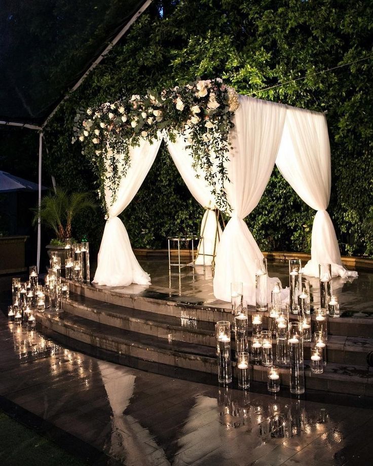 an outdoor wedding setup with white drapes and flowers on the stage, surrounded by candles