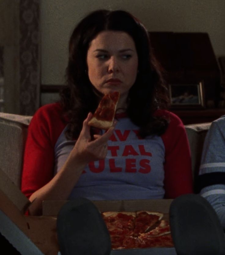 two people sitting on a couch eating pizza and looking at each other's eyes