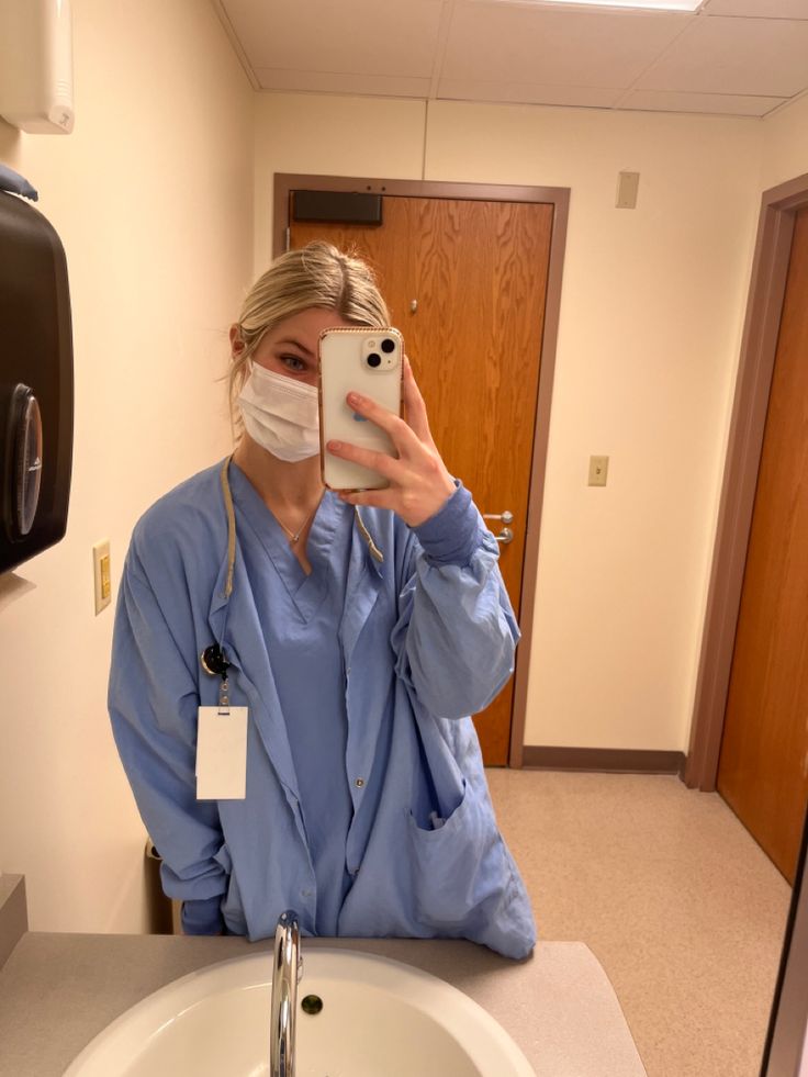 a woman in scrubs is taking a selfie with her cell phone while wearing a face mask