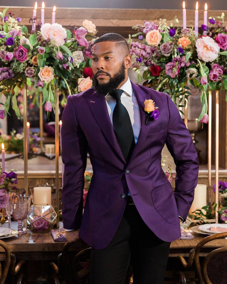 a man in a purple suit standing next to a table with candles and flowers on it